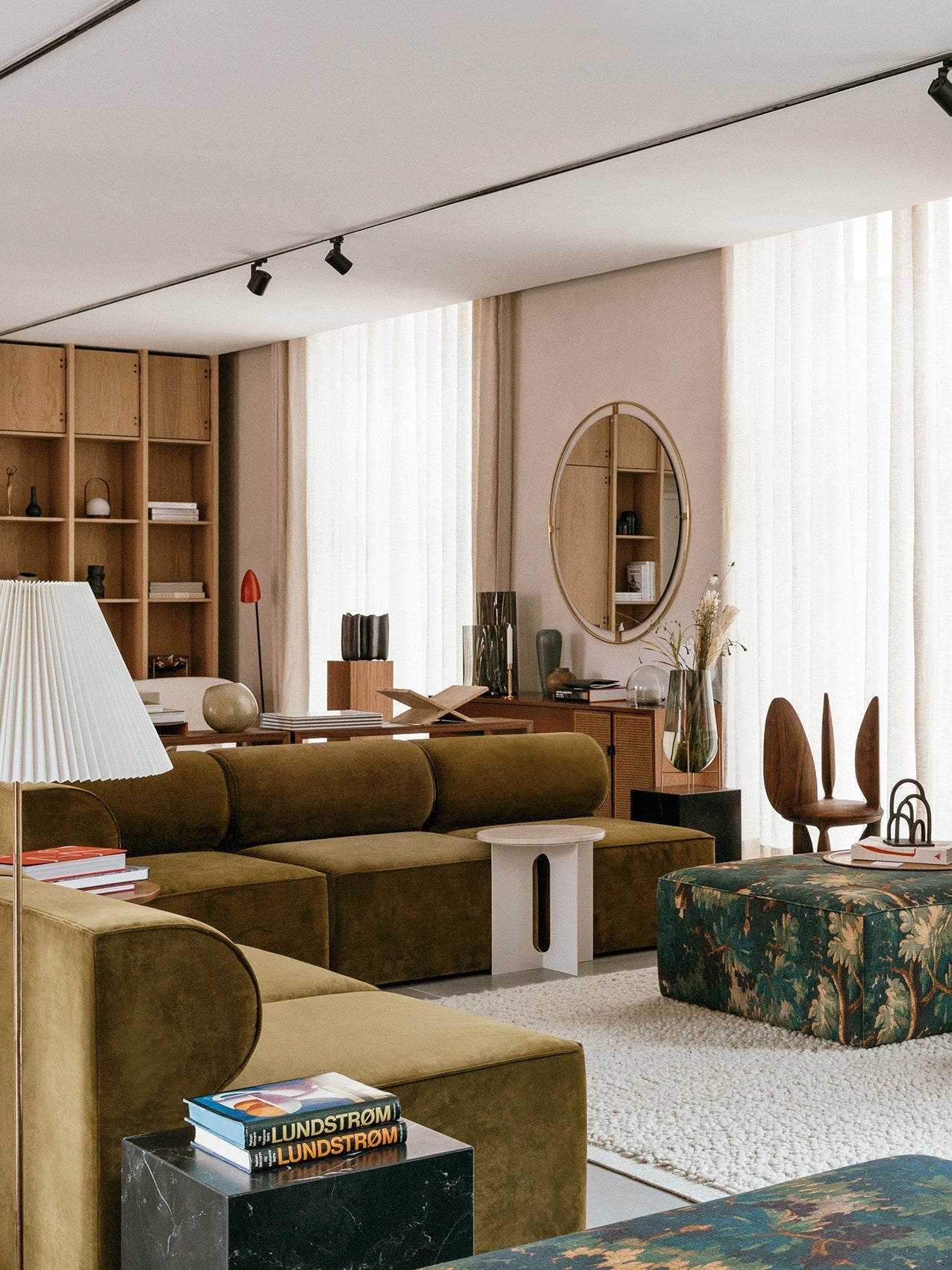 A sunlit living room with an olive green Eave Modular Sofa, a patterned ottoman, wooden shelves, and a large round mirror.