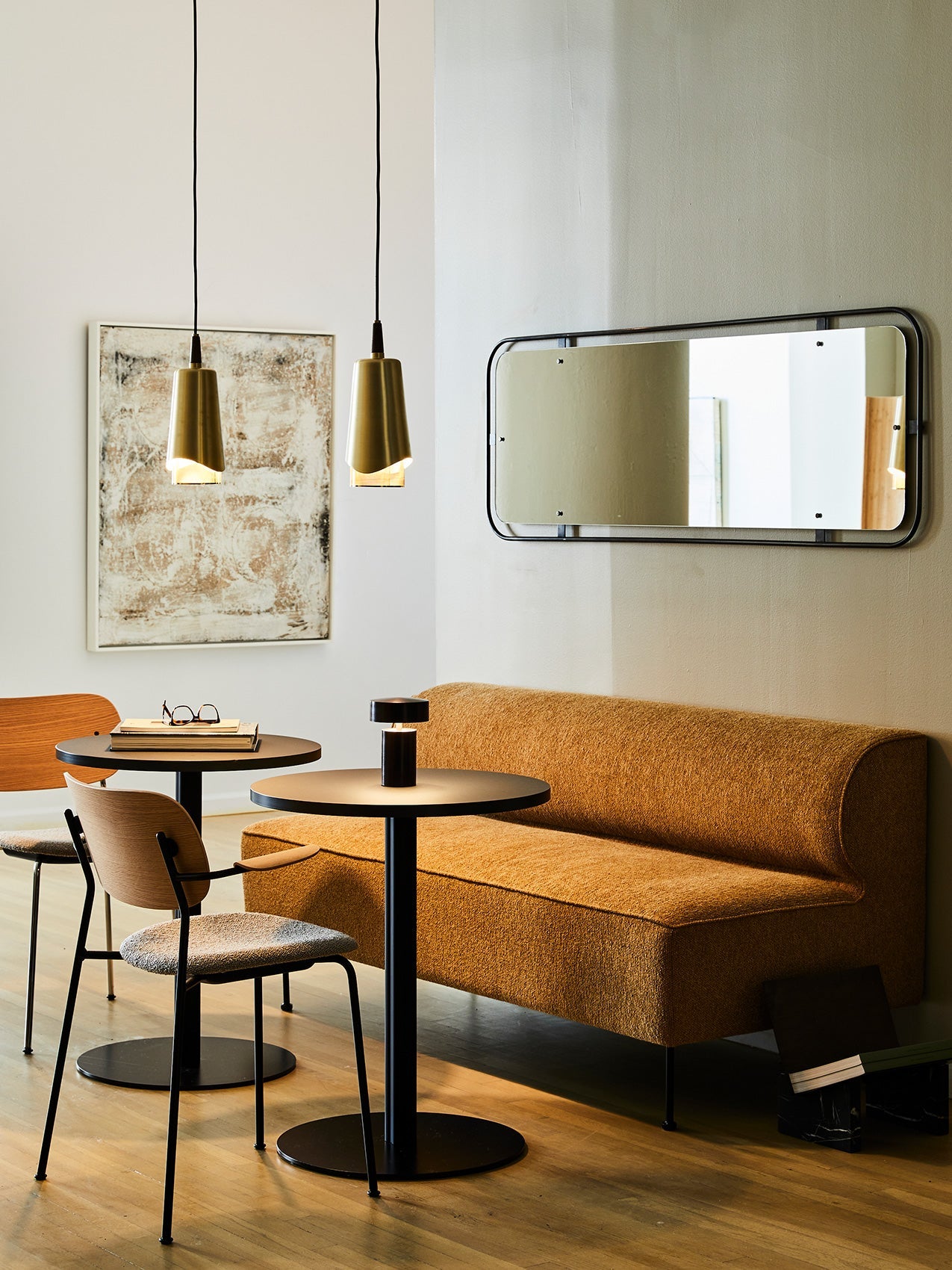 A dining corner with an orange Eave Dining Sofa, two black tables with Co Chairs, a Column Table Lamp, and brass pendants.