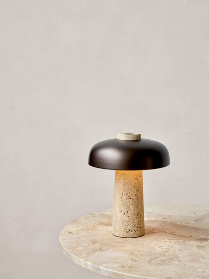 The Reverse Table Lamp, with a dark shade and glowing travertine base, sits on the marble top of a Harbour Column Table.
