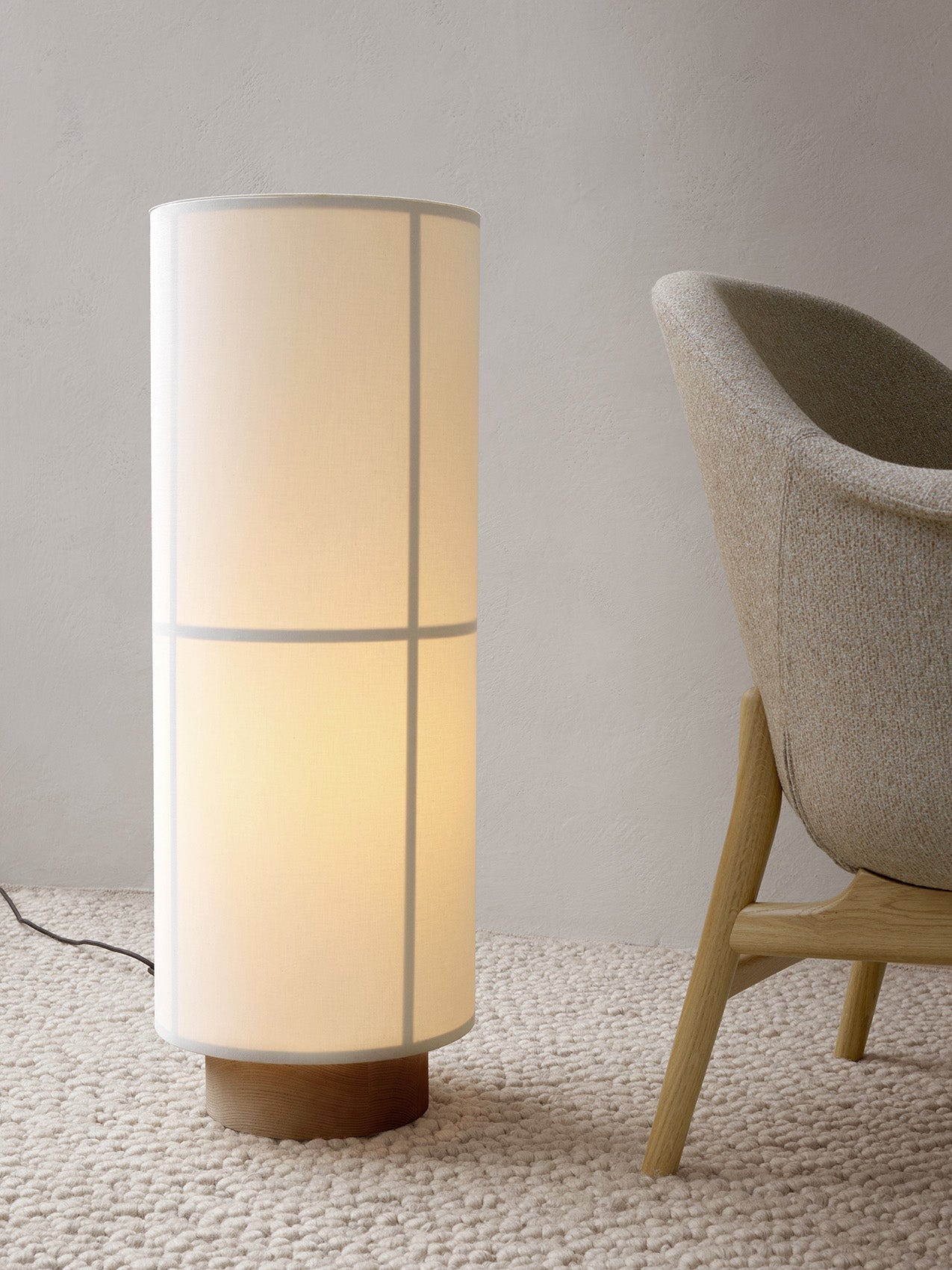 A lit Hashira Floor Lamp with a linen shade and wood base sits on a cream Gravel Rug next to a Harbour Lounge Chair.