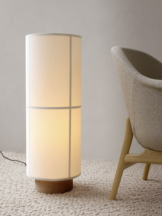 A lit Hashira Floor Lamp with a linen shade and wood base sits on a cream Gravel Rug next to a Harbour Lounge Chair.