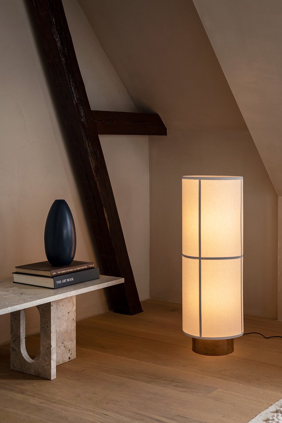 The Hashira Floor Lamp lights a cozy attic room, standing on a wooden floor beside a low stone table with books and a vase.