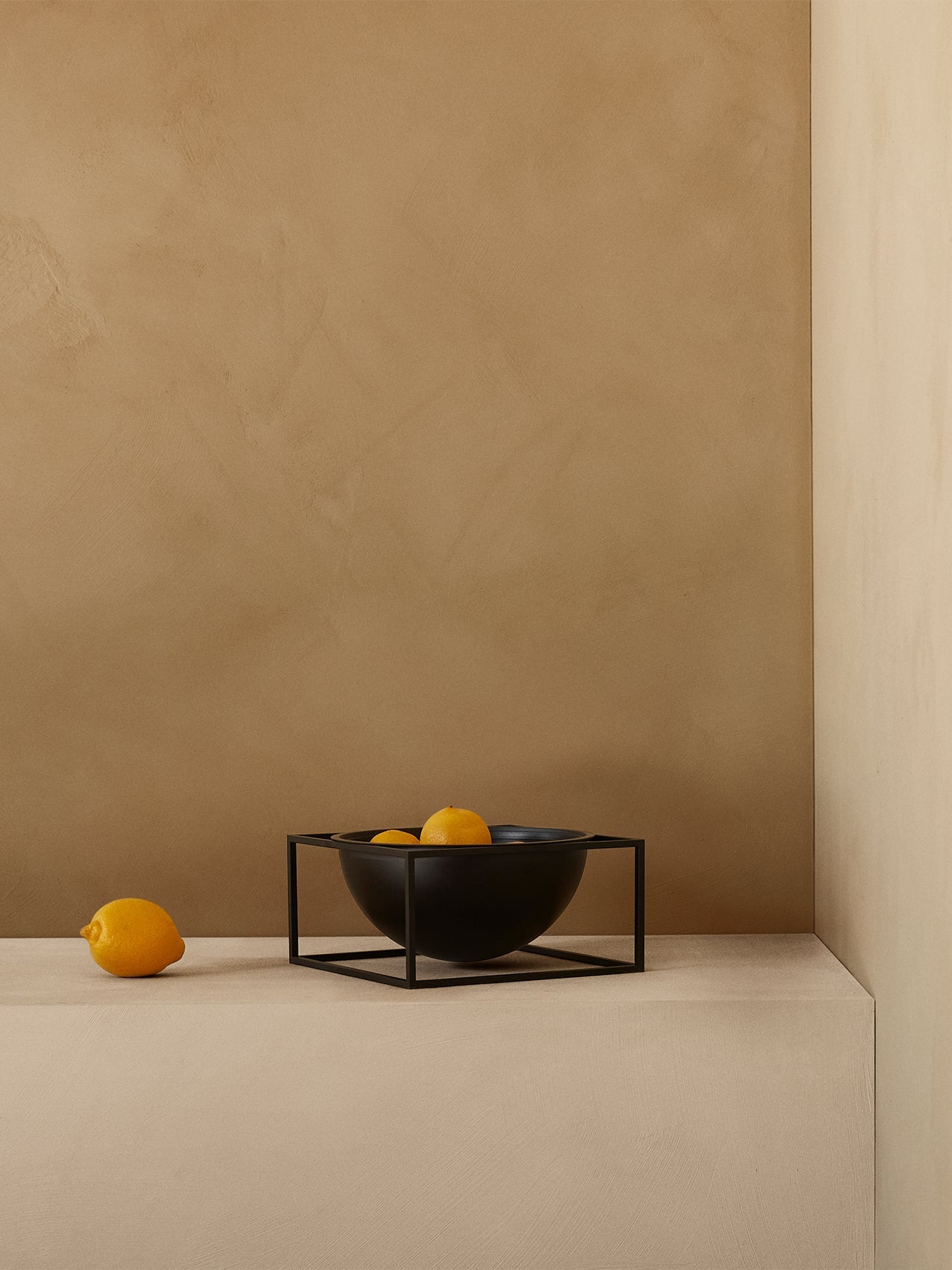 The black Kubus Bowl Centerpiece, featuring a geometric metal frame, holding lemons on a beige shelf against a beige wall.