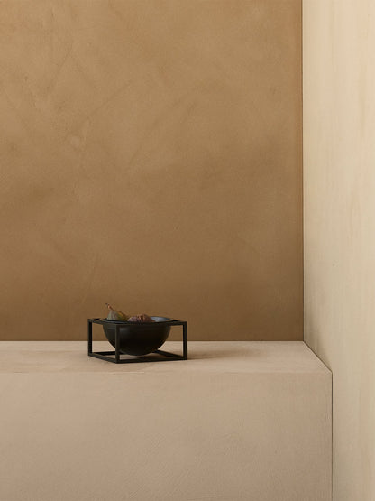 The black metal Kubus Bowl Centerpiece holding figs, styled on a beige surface in a minimalist interior setting.