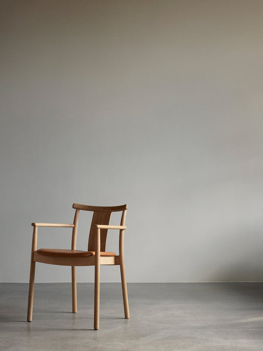 A Merkur Dining Chair with Armrests in natural oak with a tan leather seat, standing in a minimalist room with a grey wall.