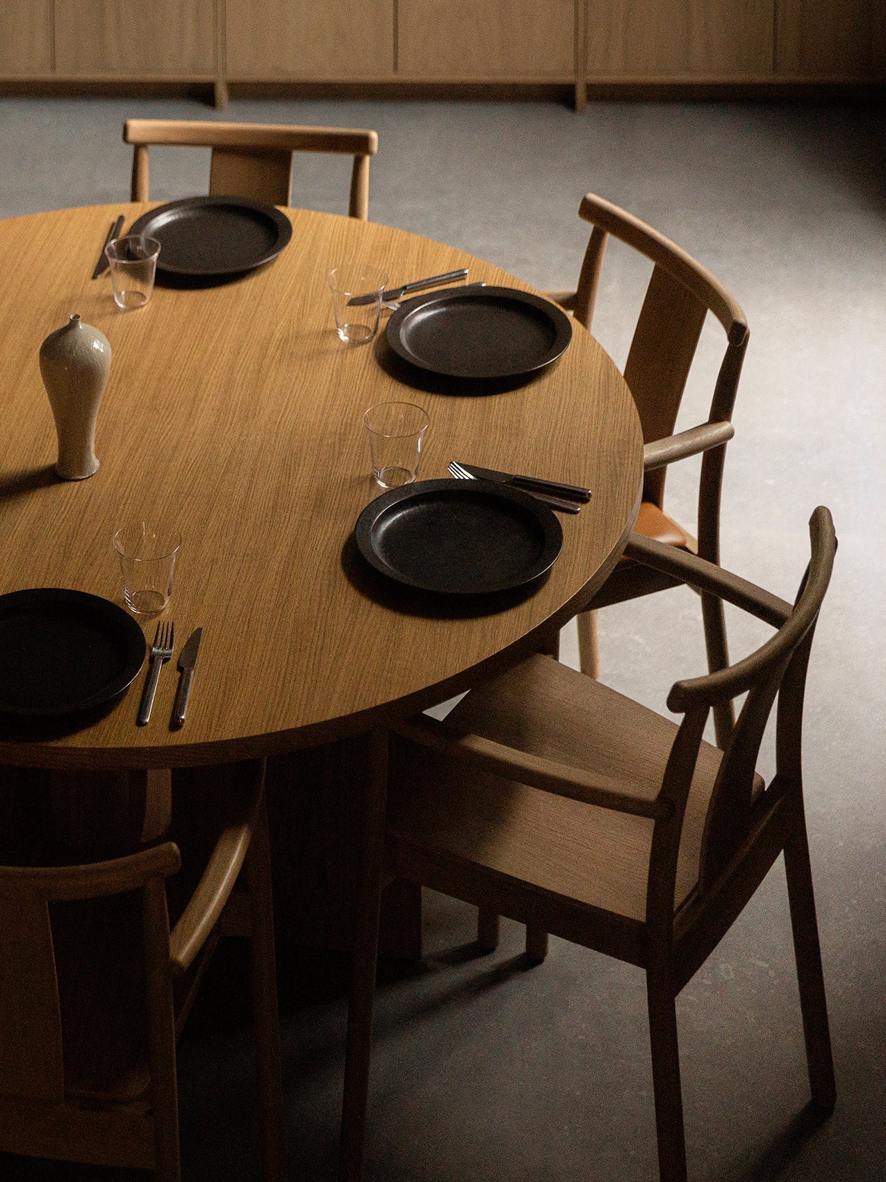 High-angle view of four oak Merkur Dining Chairs with armrests surrounding a round oak Androgyne Dining Table set for a meal.