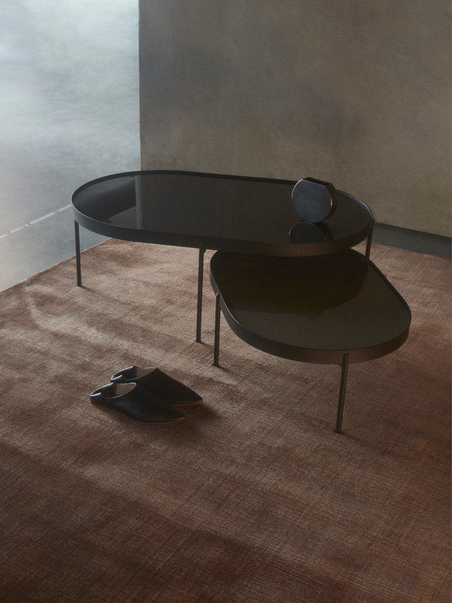 A large and a small NoNo Table with reflective brown glass tops and black frames, set on a brown rug in a minimalist room.