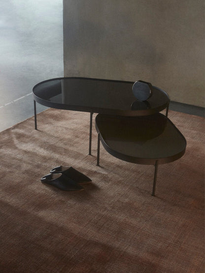 A large and a small NoNo Table with reflective brown glass tops and black frames, set on a brown rug in a minimalist room.
