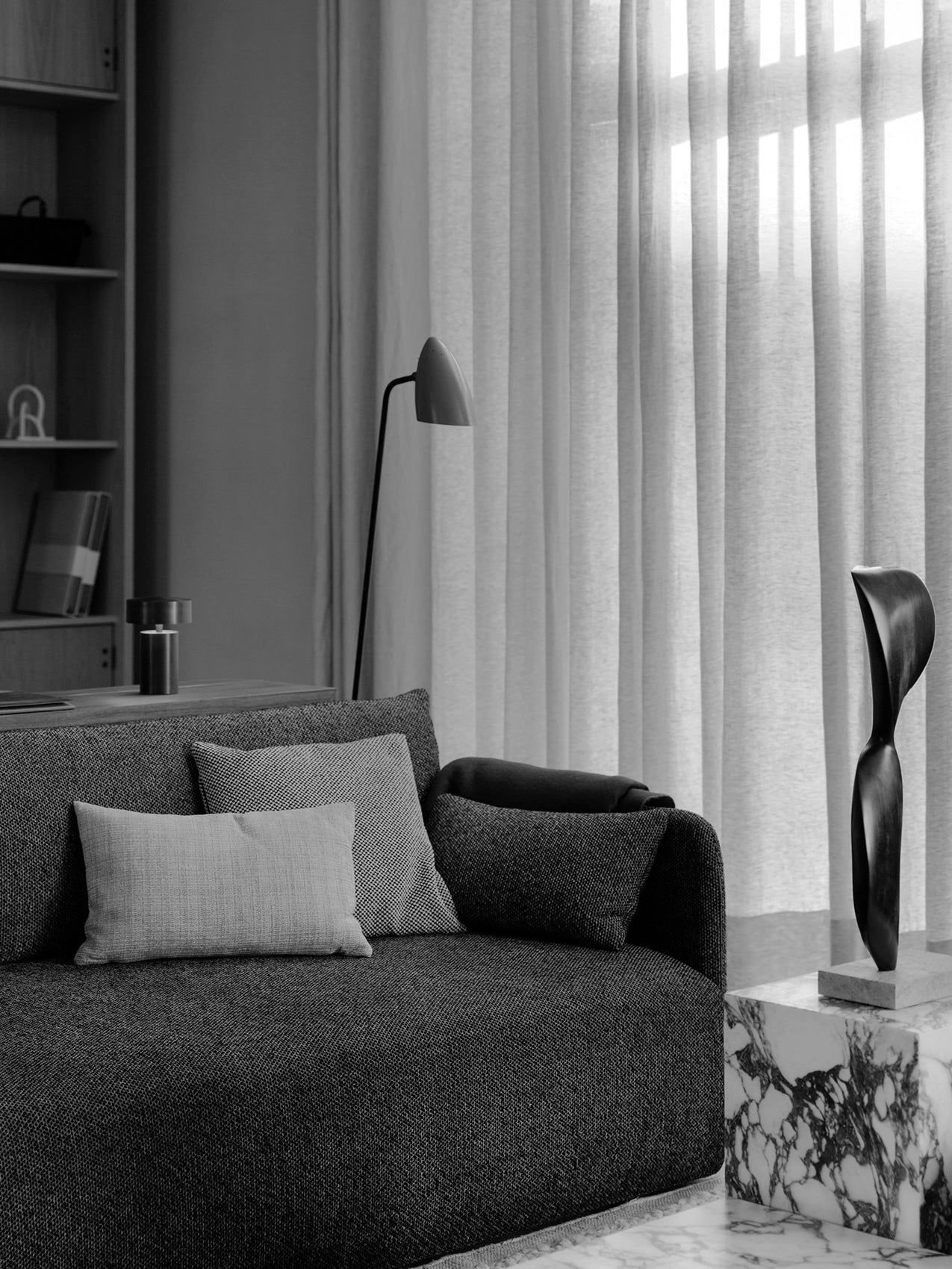 A black and white shot of the Offset Sofa in a living room setting, beside a marble Plinth and in front of a floor lamp.