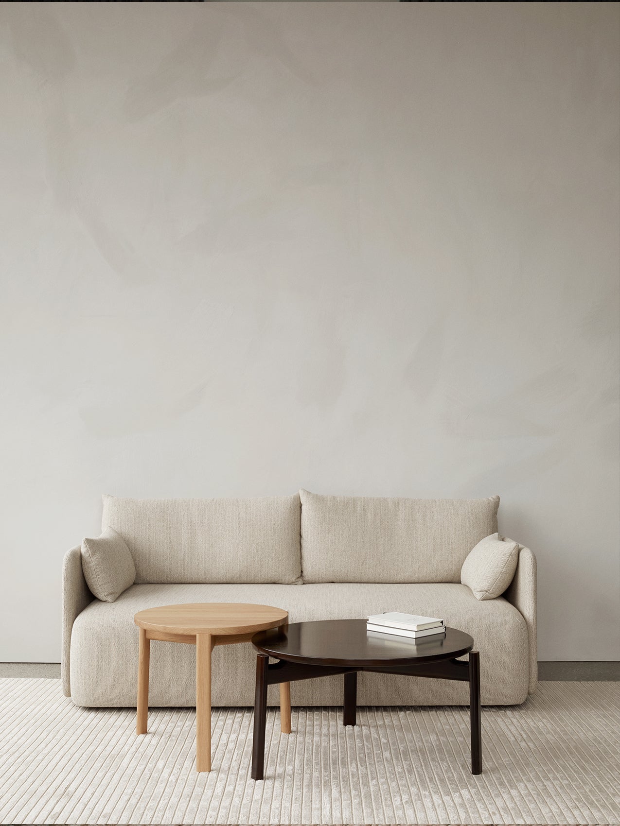 In a minimalist room, a beige Offset Sofa is styled with two Passage Lounge Tables, in oak and dark wood, on a textured rug.