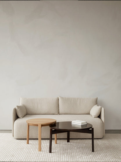 In a minimalist room, a beige Offset Sofa is styled with two Passage Lounge Tables, in oak and dark wood, on a textured rug.
