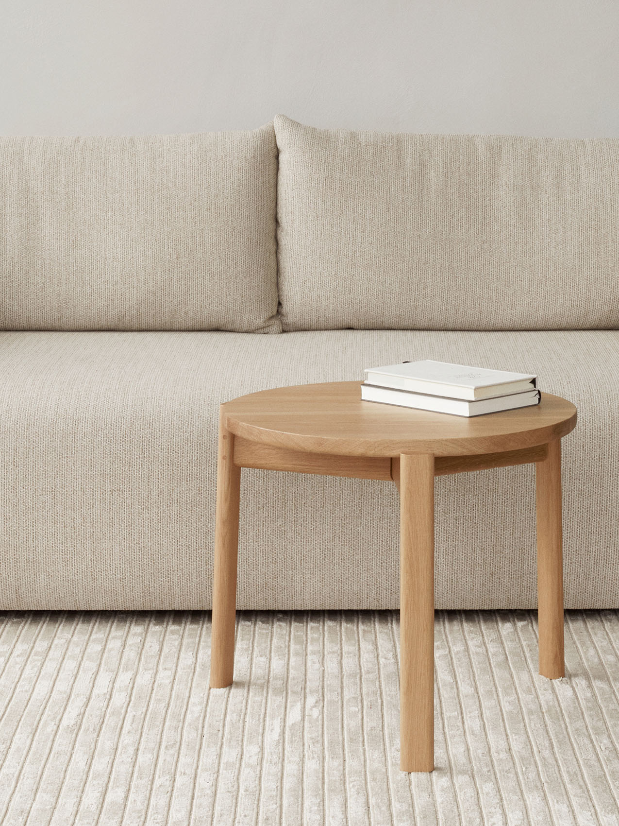 A close-up of a round oak Passage Lounge Table with two white books, set against a beige Offset Sofa and on a Houkime Rug.