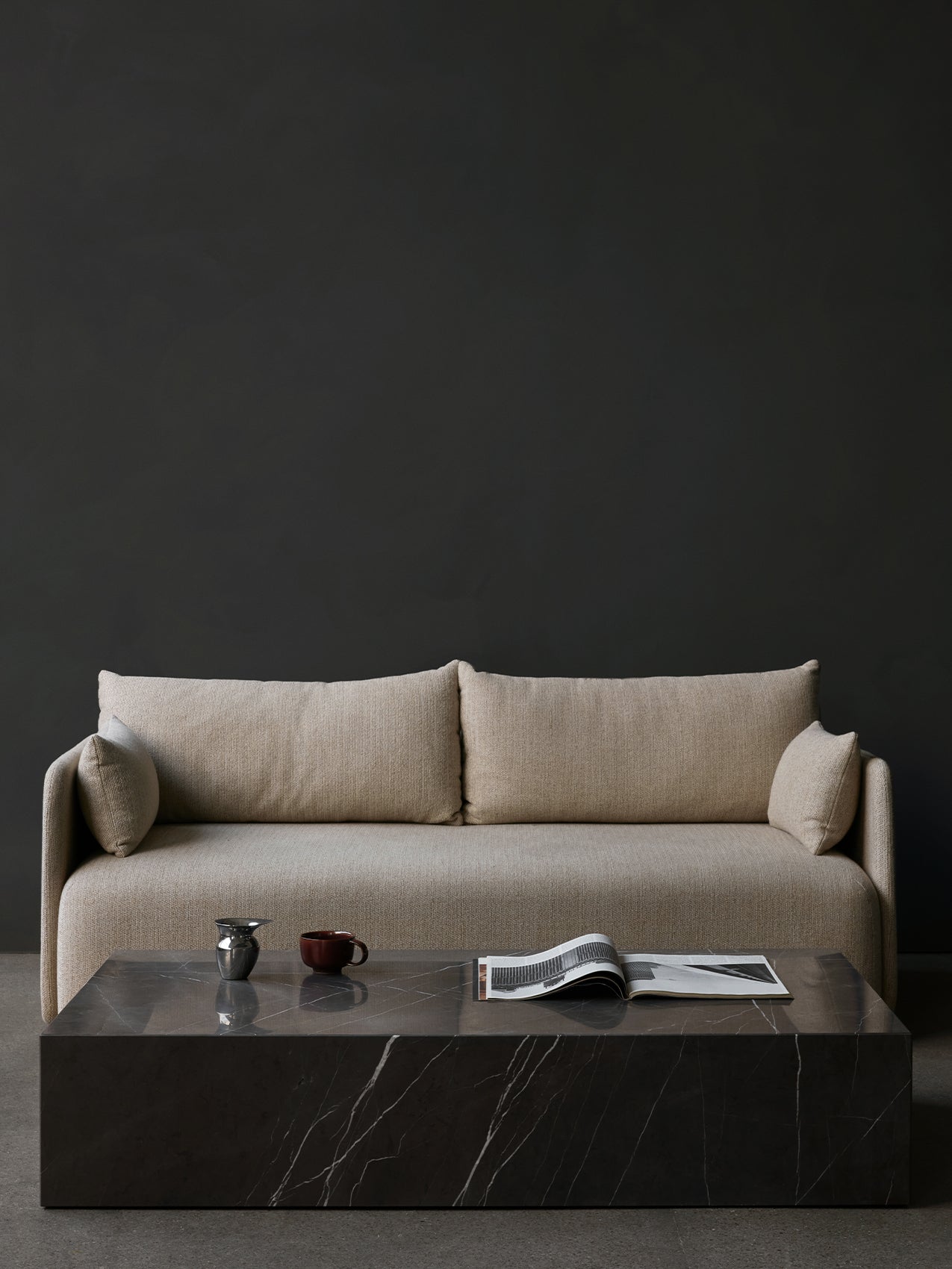 Front view of the beige Offset Sofa behind a black marble Plinth Grand coffee table with an Etruscan Jug against a dark wall.