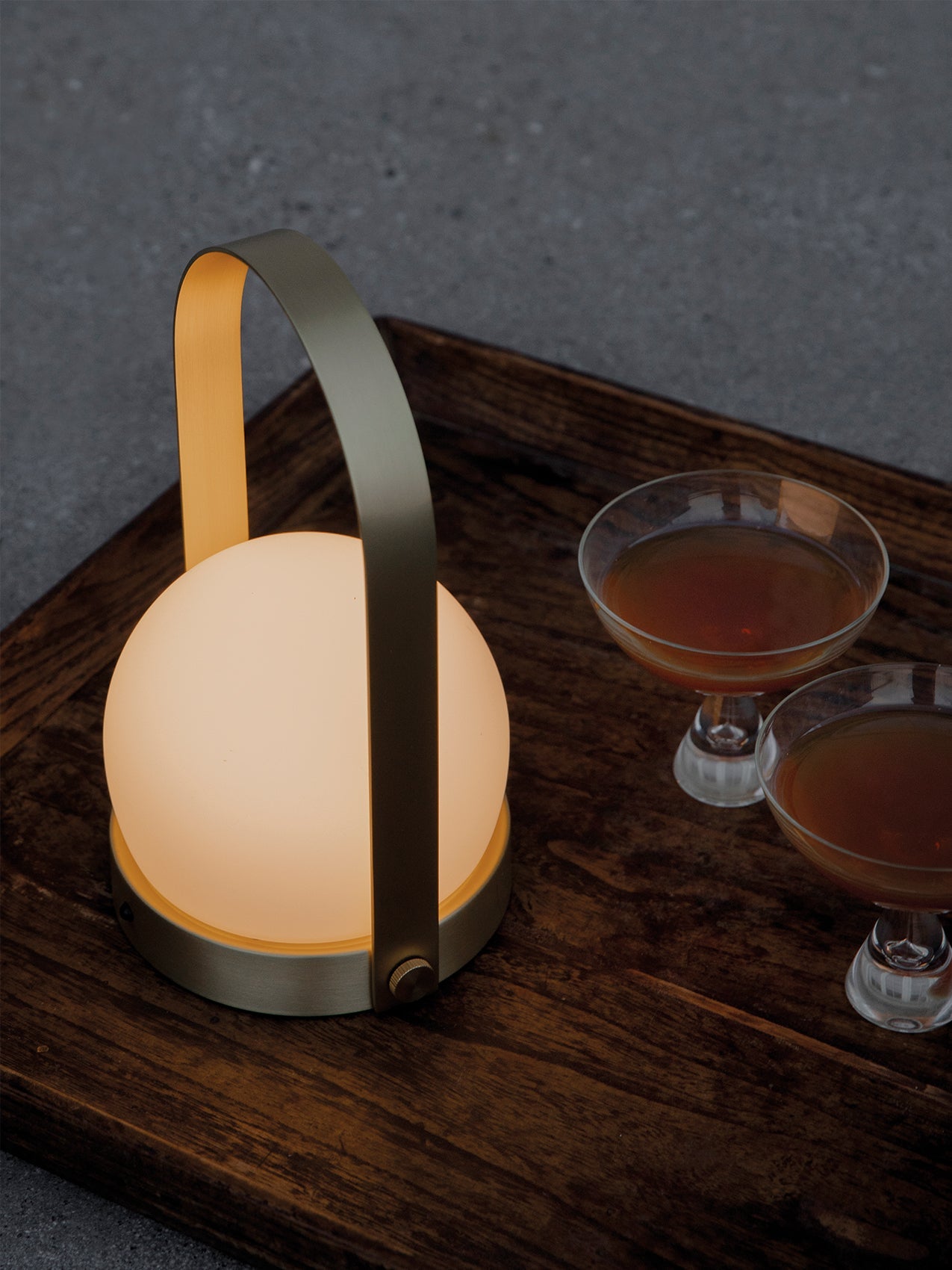 The glowing Carrie Table Lamp in Brass sits on a dark wooden tray, illuminating two coupe glasses filled with cocktails.