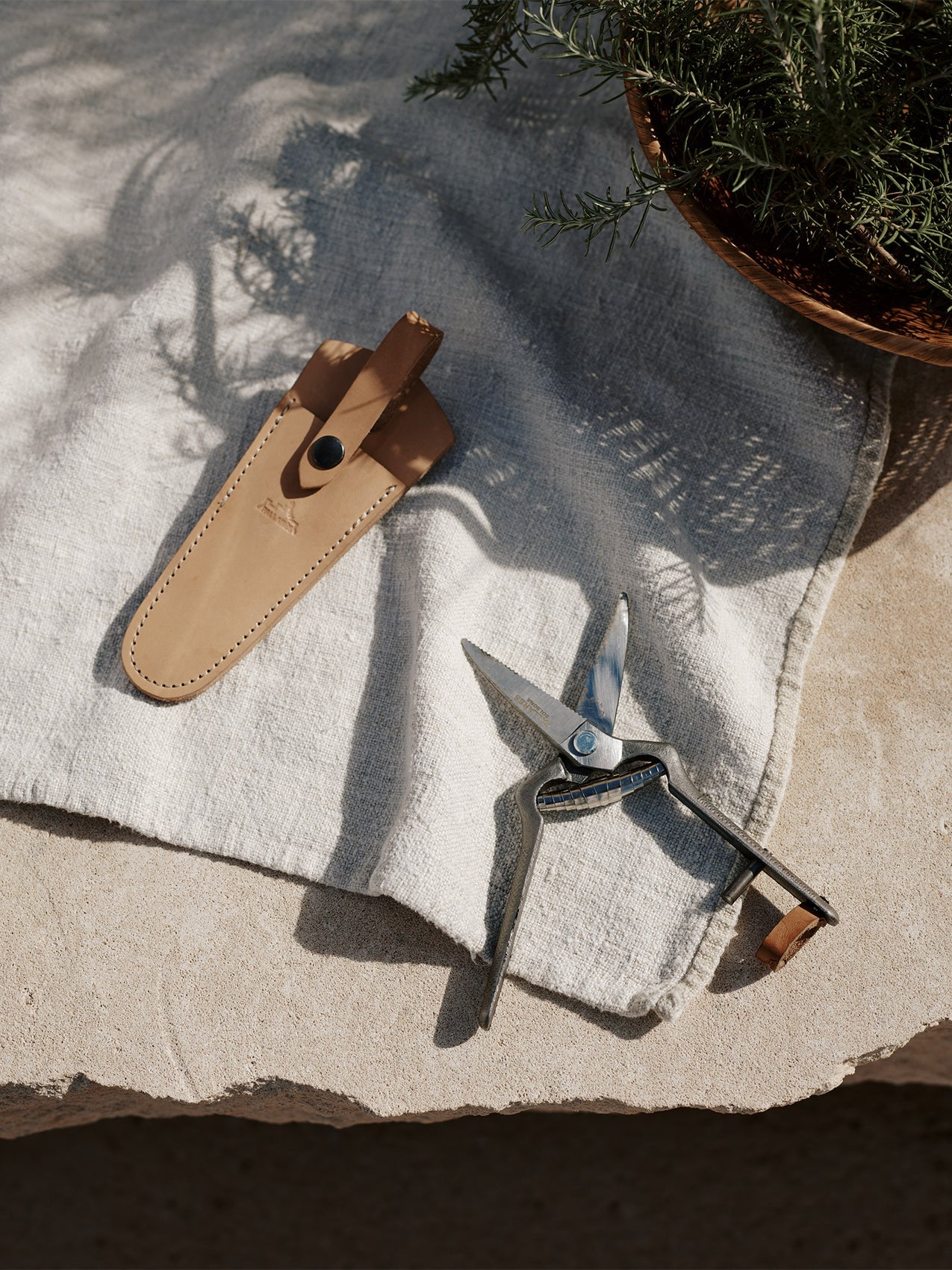 The Pallares Plant Pruner and its tan leather sheath resting on a light linen cloth on a stone surface by a bowl of herbs.