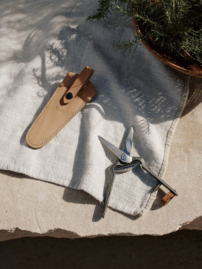 The Pallares Plant Pruner and its tan leather sheath resting on a light linen cloth on a stone surface by a bowl of herbs.
