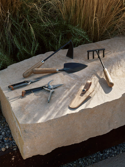 Pallares Plant Set: hand hoe, trowel, rake, pruning shears, snips, and a leather sheath arranged on a large garden stone.