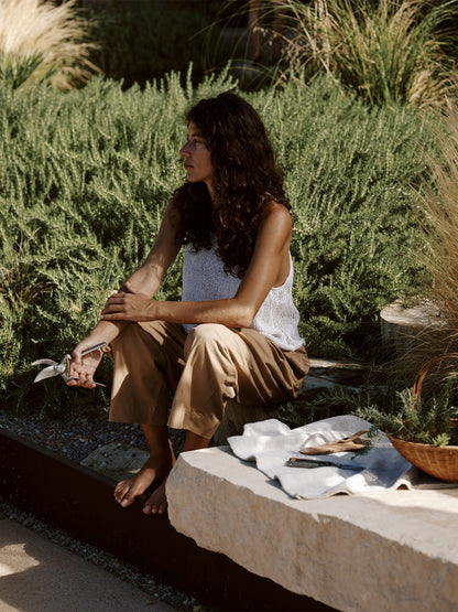 A woman with long curly hair sits barefoot on stone steps in a sunny garden, holding the silver Pallares Plant Shears.