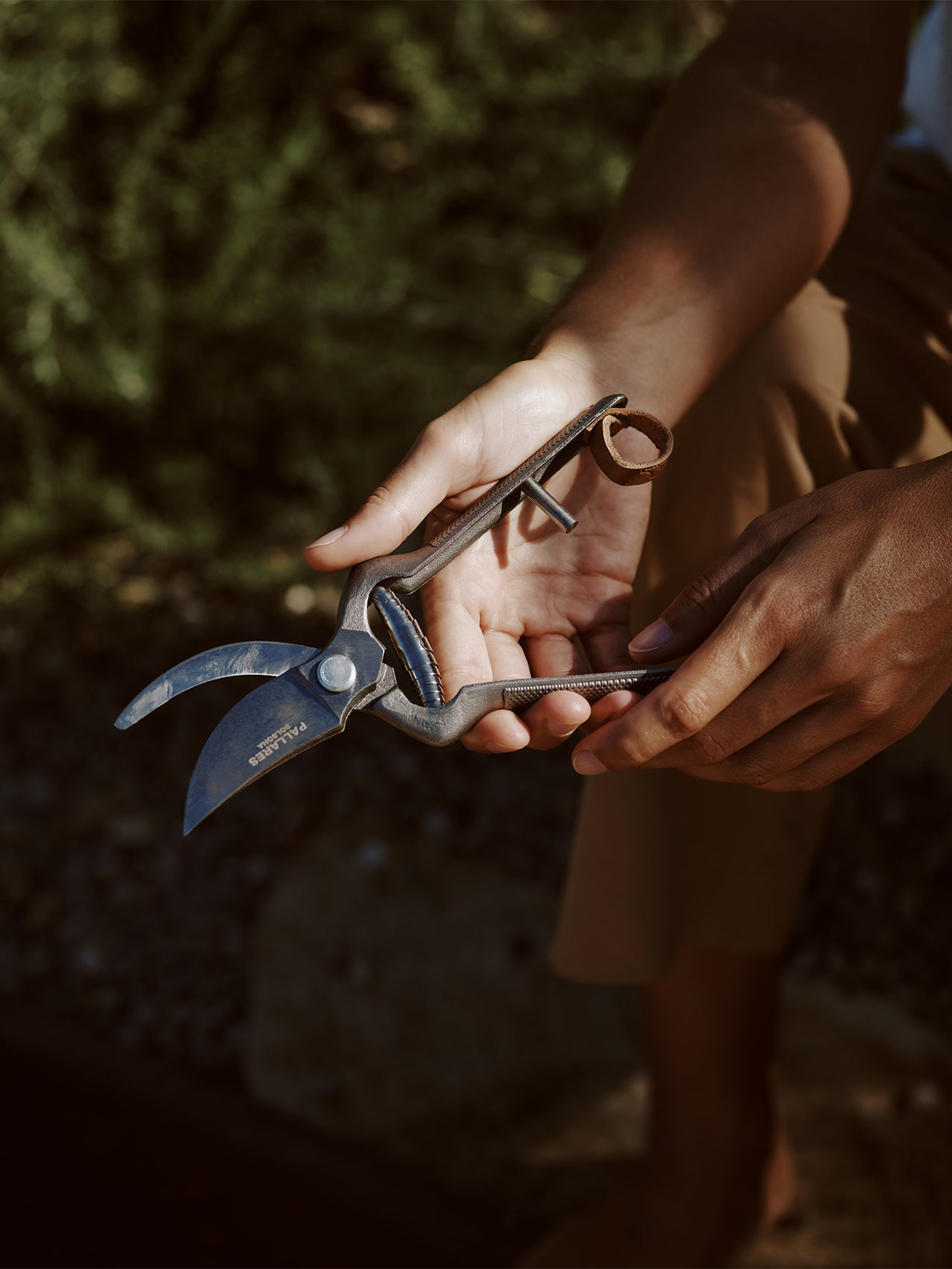 A person's hands holding dark metal Pallares Plant Shears in a sunlit garden, showing textured handles and curved blades.