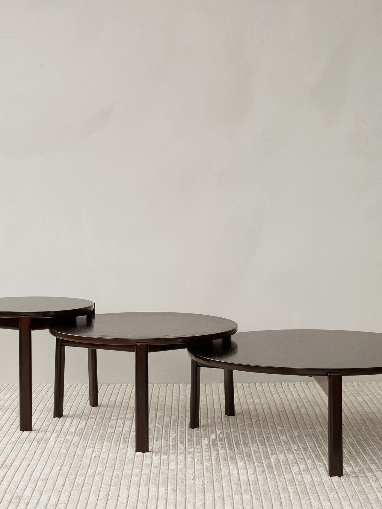 Three dark wood Passage Lounge Tables of different sizes arranged in a nesting formation on a light grey Houkime rug.