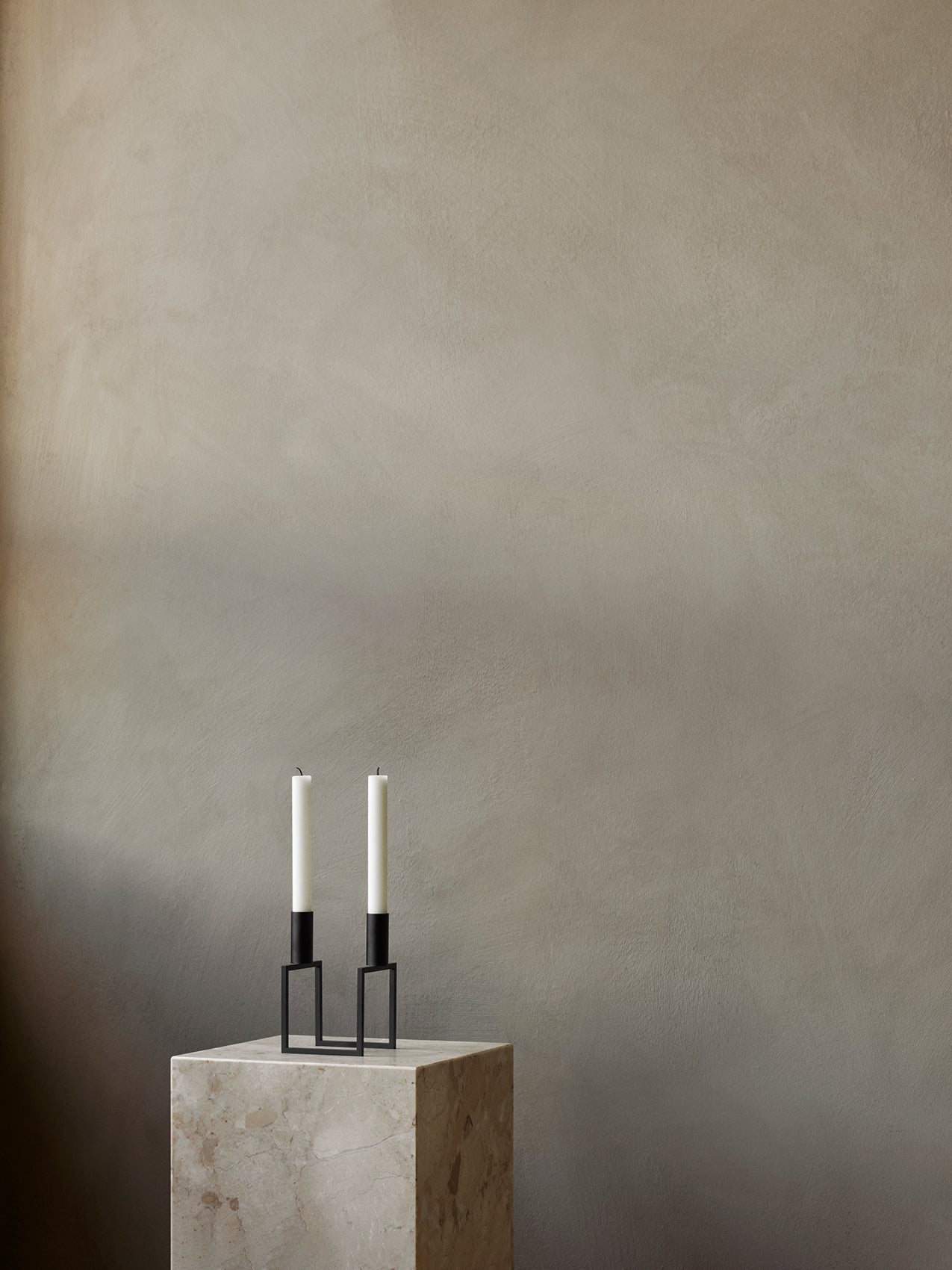 A black Kubus Line Candle Holder with two white candles on a marble Plinth Pedestal against a muted, textured beige wall.