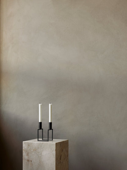 A black Kubus Line Candle Holder with two white candles on a marble Plinth Pedestal against a muted, textured beige wall.