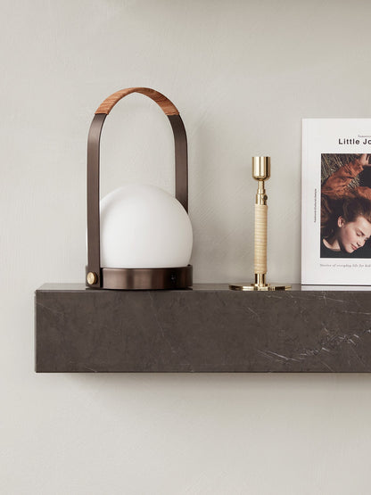 The bronzed brass Carrie Table Lamp sits on a dark grey marble Plinth Shelf next to a brass Duca Candle Holder and a book.