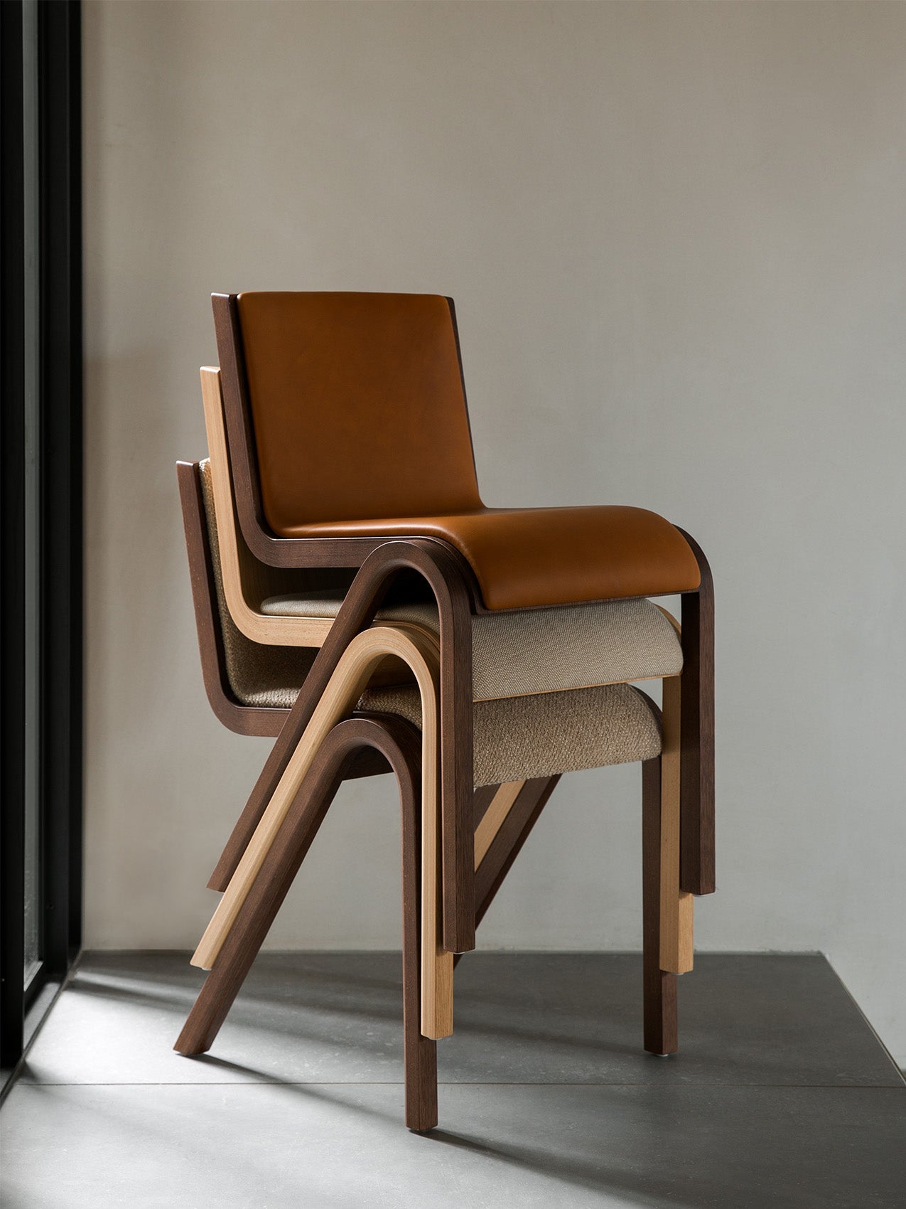 A stack of three upholstered Ready Dining Chairs in different finishes: dark wood and tan leather, and oak with beige fabric.