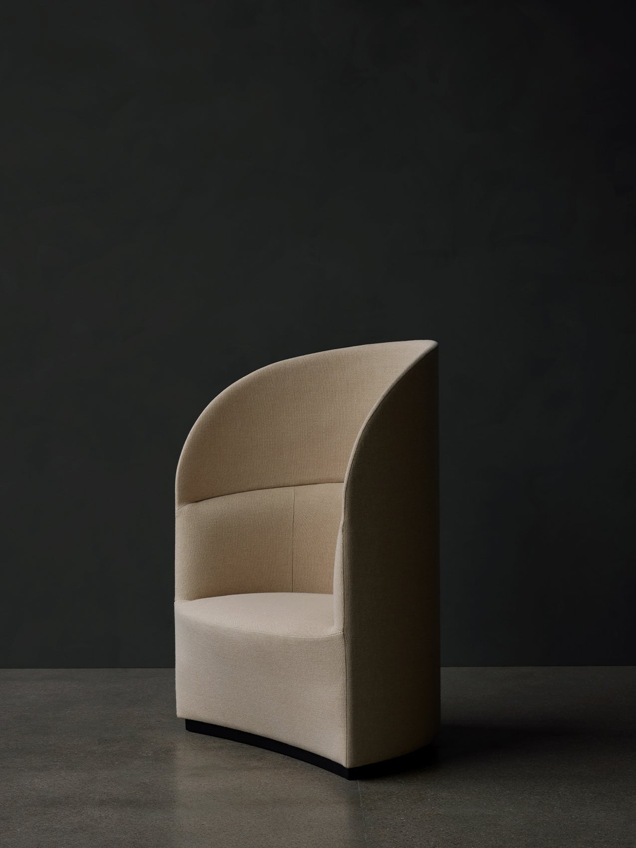 Sculptural Tearoom Lounge Chair, High Back in light beige fabric, with a high, enveloping back, on a dark concrete floor.