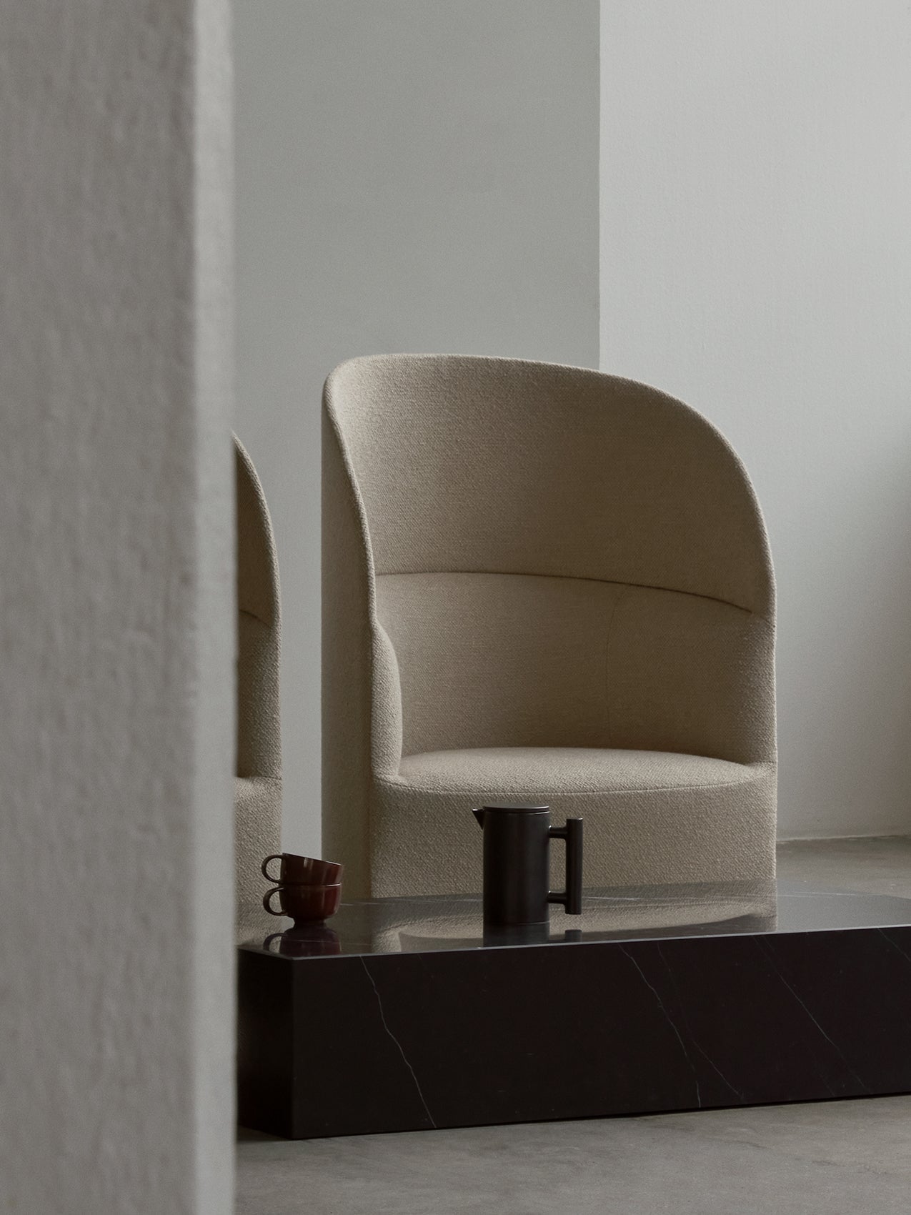 A beige Tearoom Lounge Chair, High Back, sits behind a black marble Plinth table with a Yana Brewing Pot and two cups.