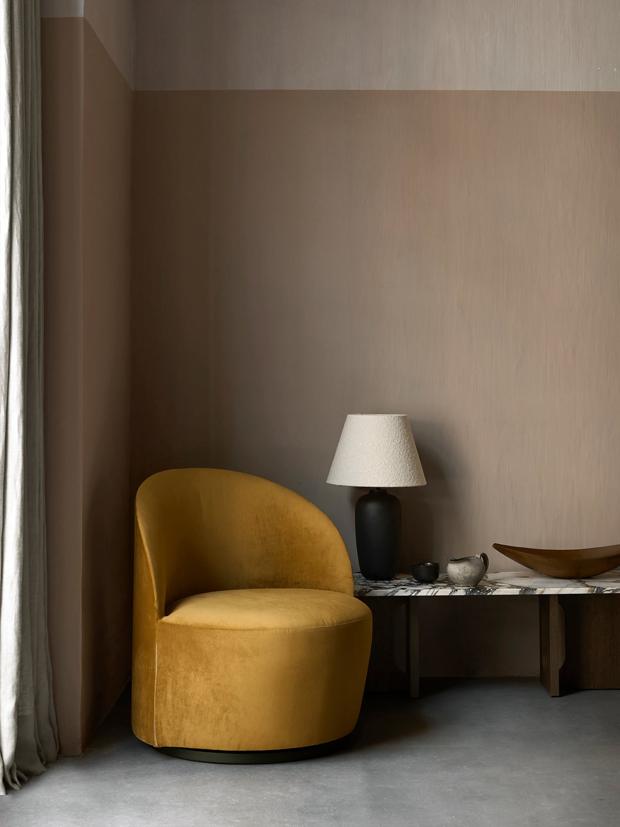 A yellow velvet Tearoom Lounge Chair in a room corner, next to a marble table with a Torso Table Lamp and decorative objects.