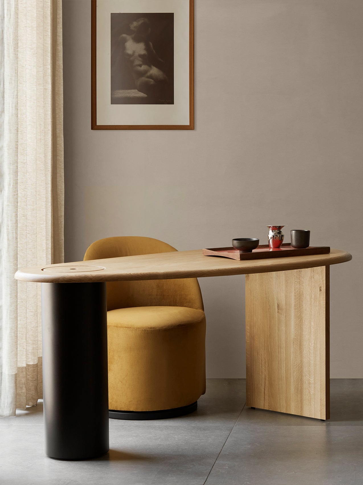 A yellow Tearoom Club Chair is tucked under the wooden Eclipse Desk in a softly lit, minimalist room with art on the wall.