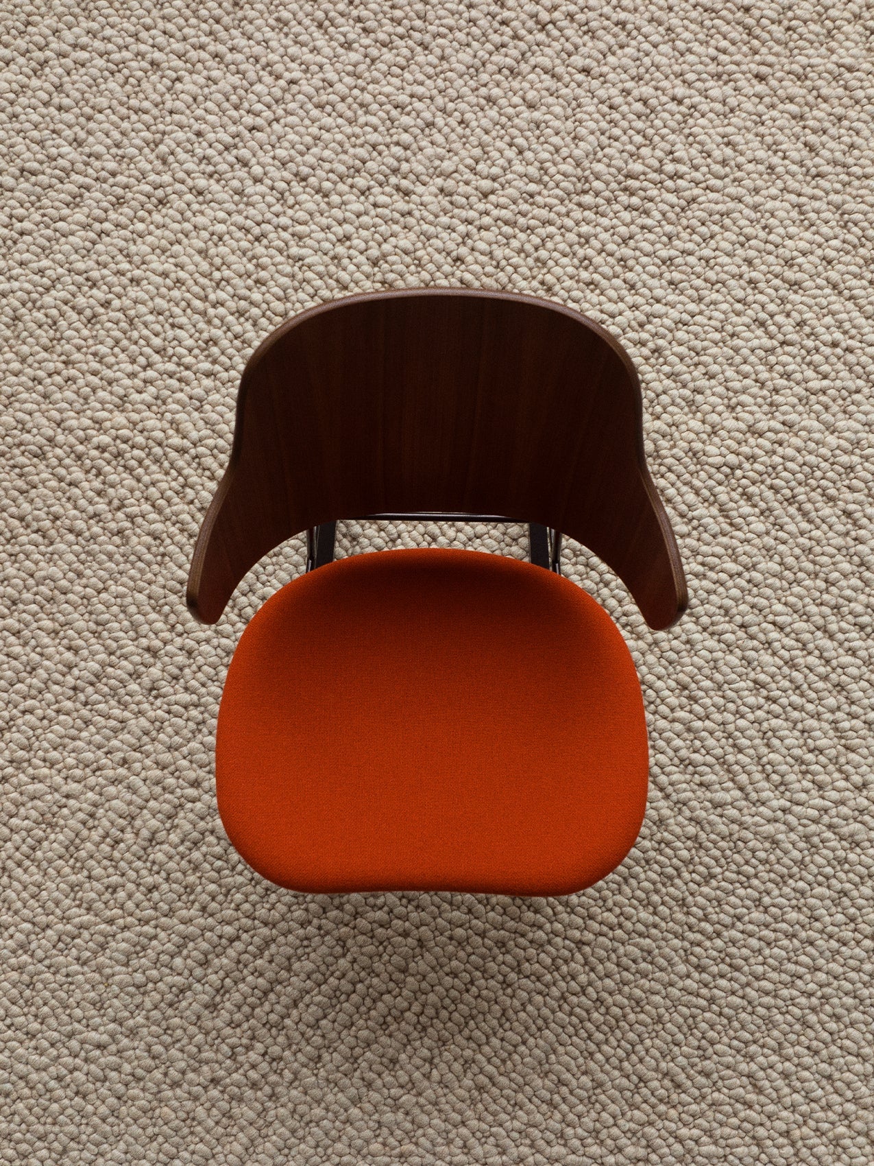 A top-down view of The Penguin Dining Chair, with a walnut backrest and orange upholstered seat, on a textured beige rug.