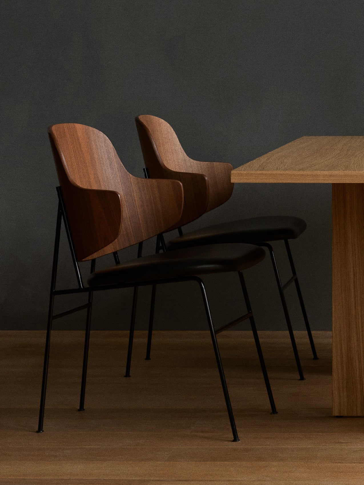 Two Penguin Dining Chairs in walnut with black seats tucked under an oak Androgyne Dining Table against a dark grey wall.