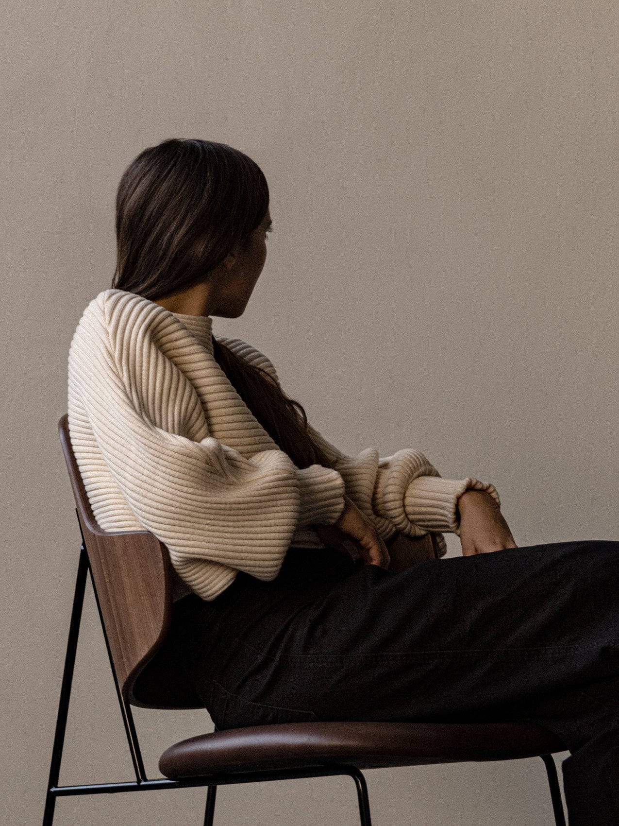 A woman wearing a cream ribbed jumper sits sideways on the dark wood Penguin Lounge Chair against a neutral background.