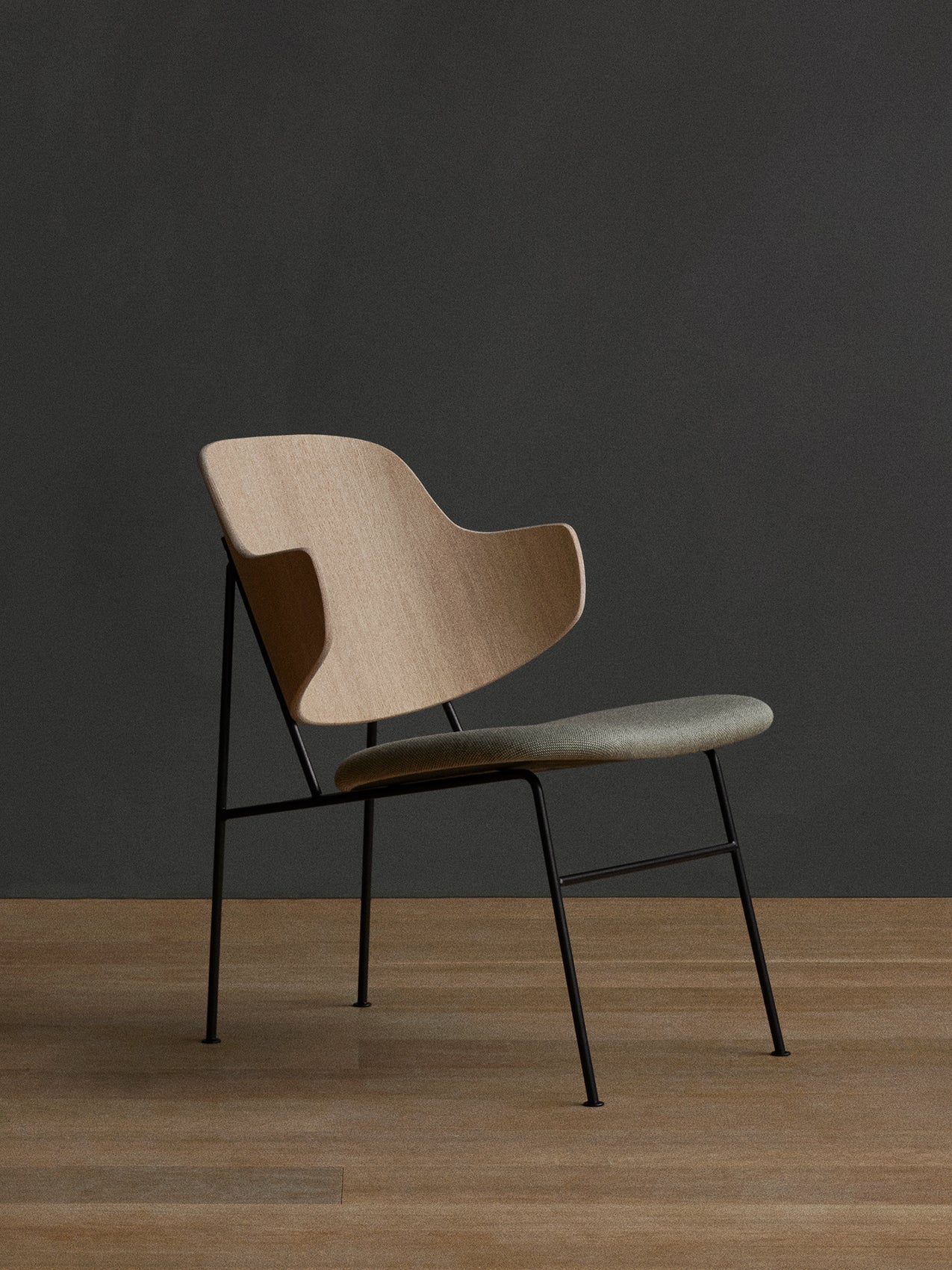 A side view of The Penguin Lounge Chair with a light wood back, grey upholstered seat, and black metal frame on a wood floor.