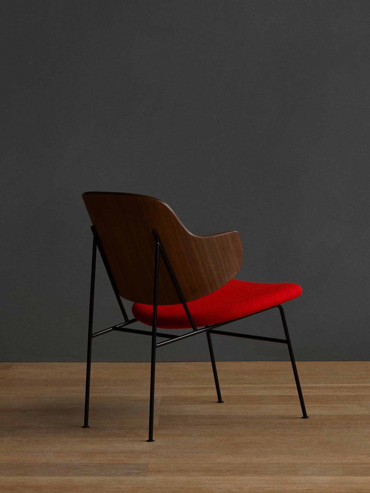 A three-quarter rear view of The Penguin Lounge Chair with a walnut veneer back, red upholstered seat, and black steel frame.