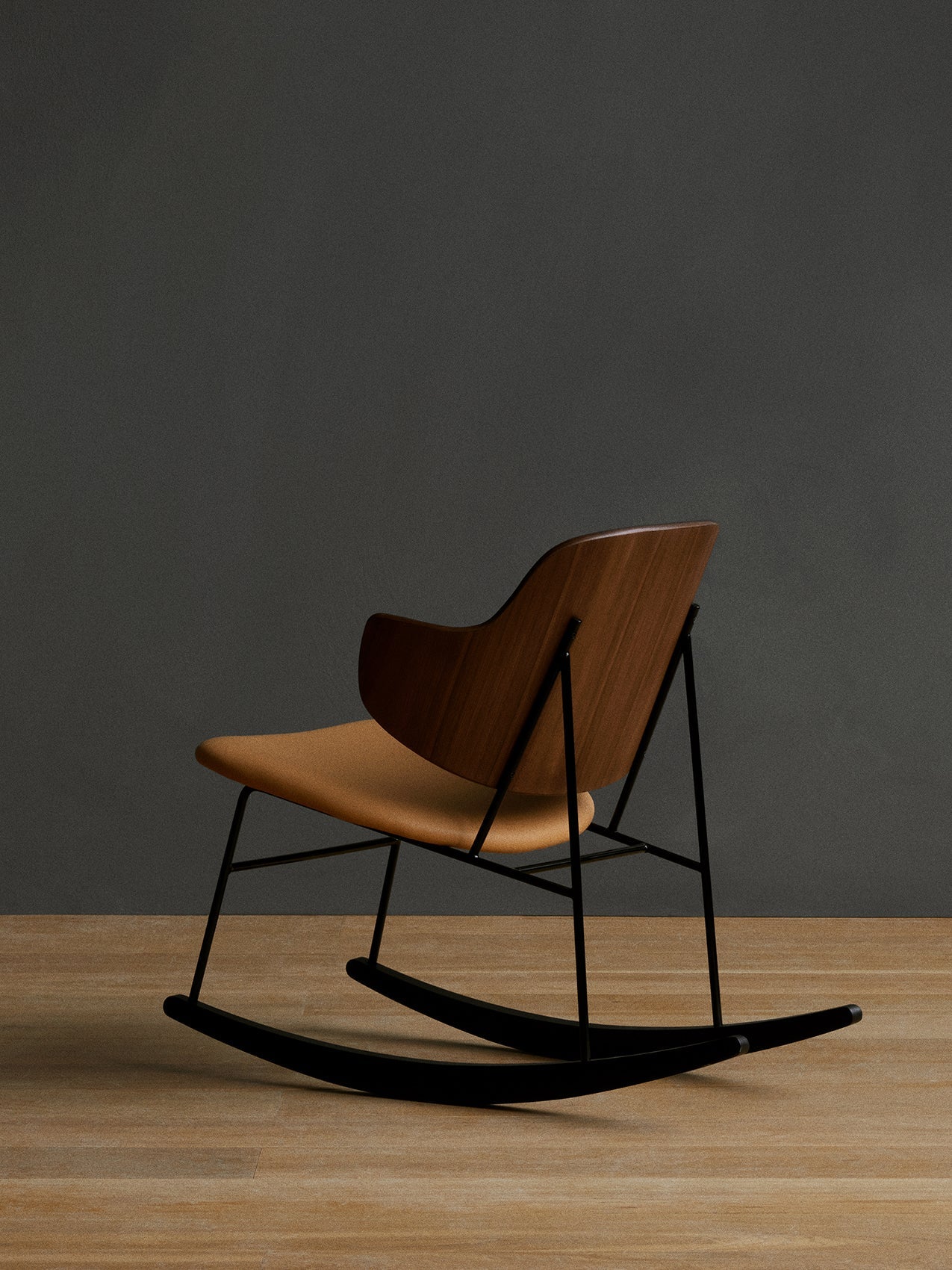Three-quarter back view of The Penguin Rocking Chair with its walnut back, tan seat and black frame on a wood floor.