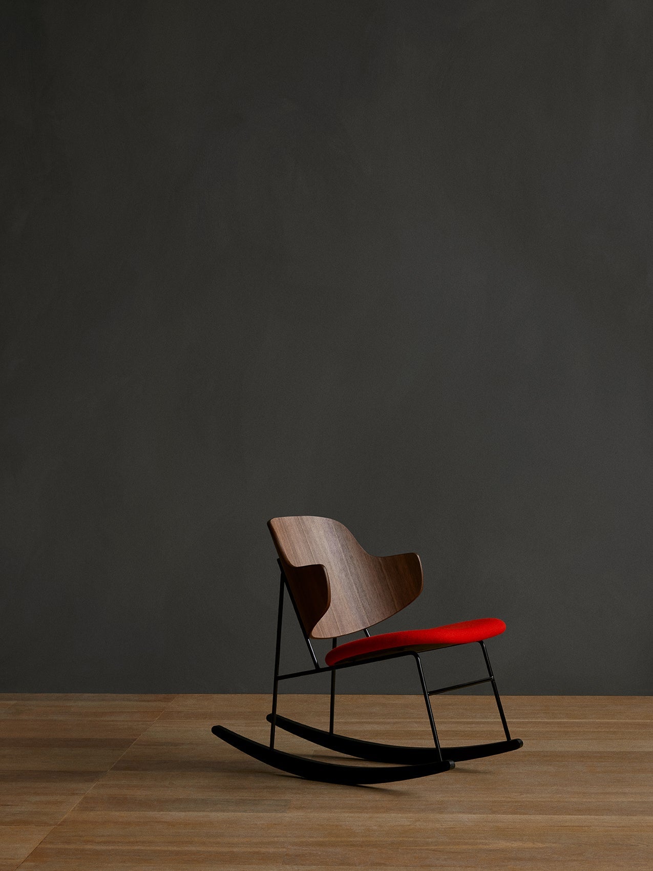 The Penguin Rocking Chair with a walnut backrest and red seat on a black frame, on a wooden floor against a dark wall.