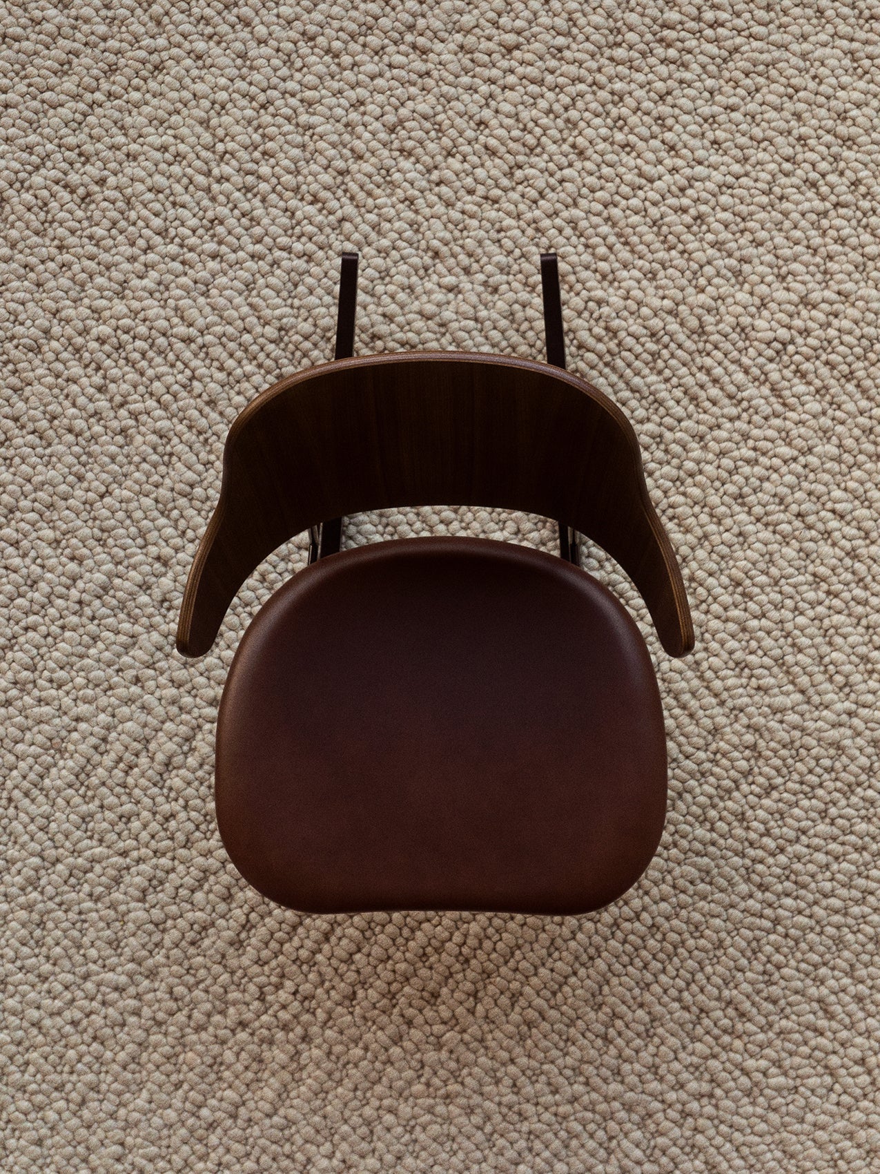 A top-down view of The Penguin Rocking Chair, with a dark wood back and a brown upholstered seat, on a textured Gravel Rug.