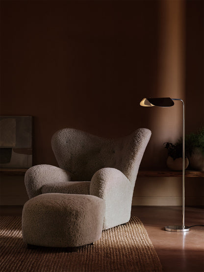 A Tired Man Lounge Chair and ottoman in soft, off-white sheepskin styled in a dark room next to an illuminated floor lamp.
