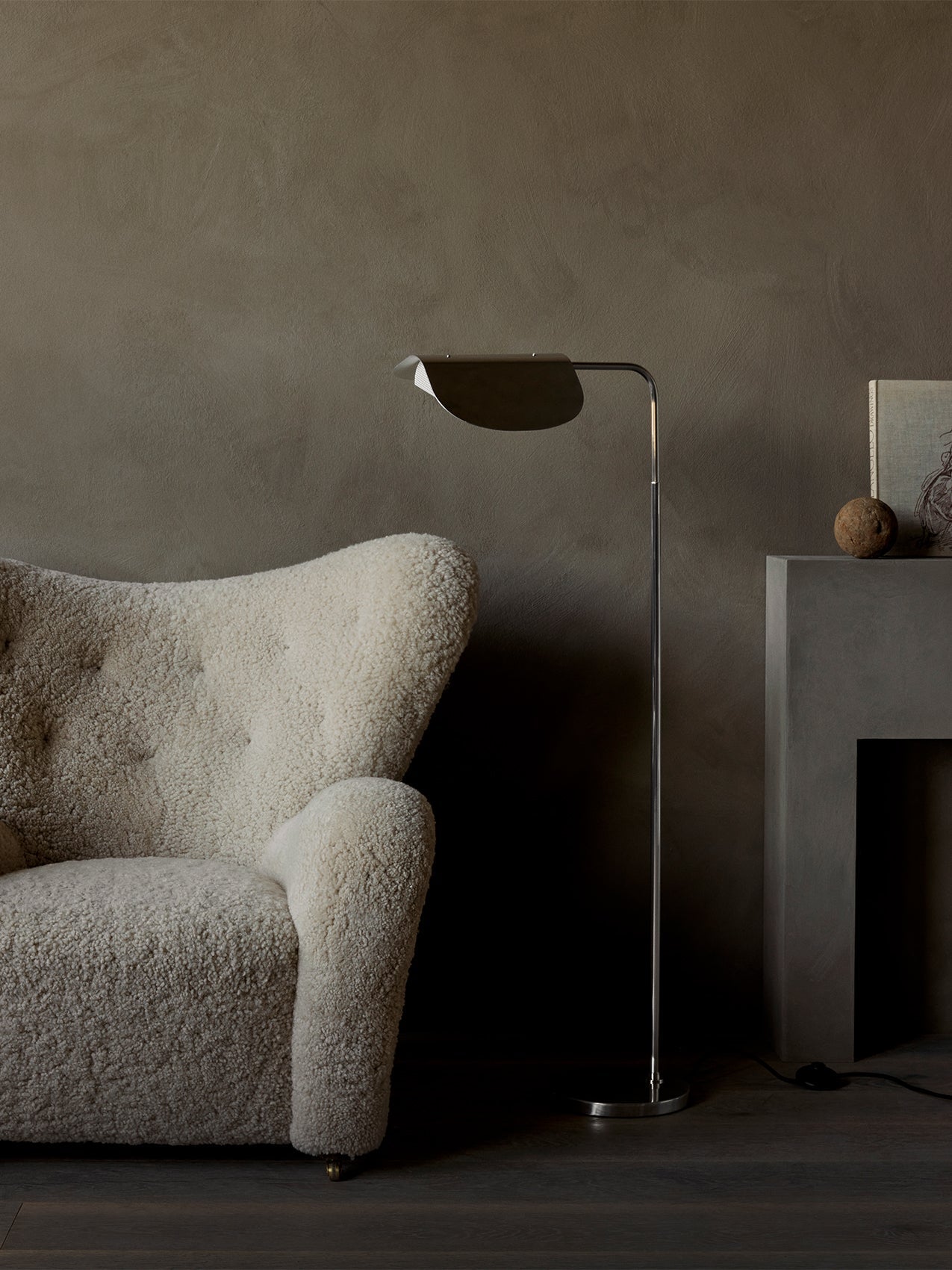 The polished aluminium Wing Floor Lamp stands in a living room beside a cream shearling armchair and a modern grey fireplace.
