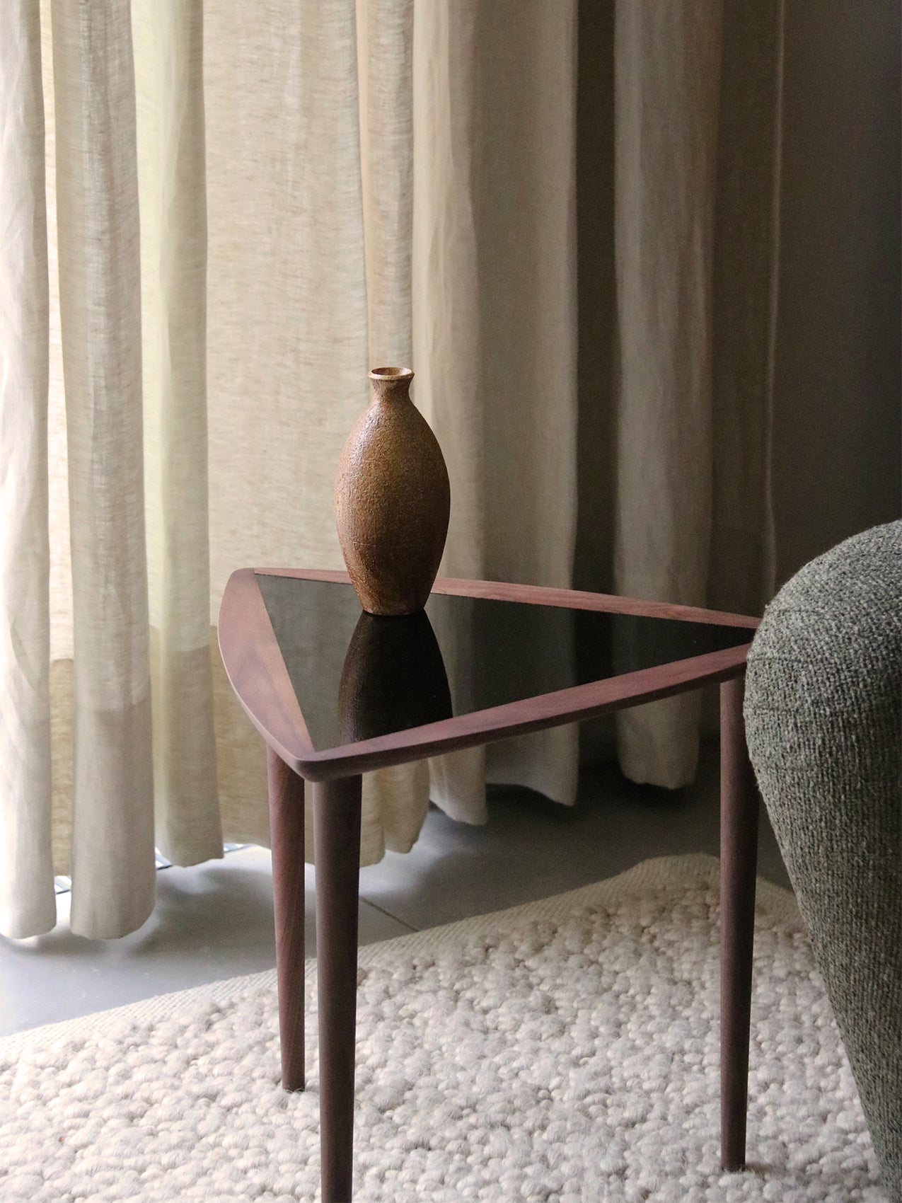 The triangular Umanoff Nesting Side Table in walnut with a black top, shown with a brown vase on a fluffy rug beside a sofa.