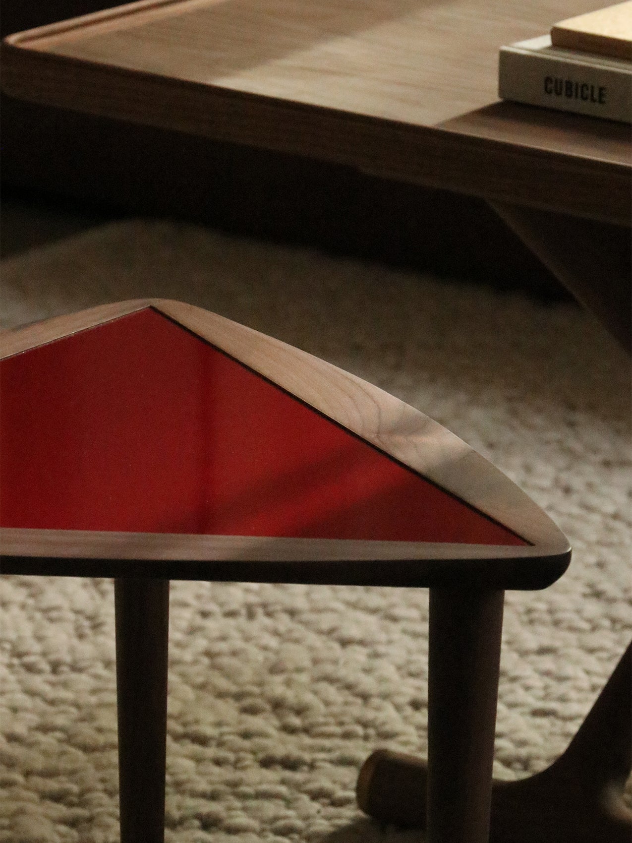 A close-up of the Umanoff Nesting Side Table, showing its triangular burgundy top and walnut frame, placed on a thick rug.