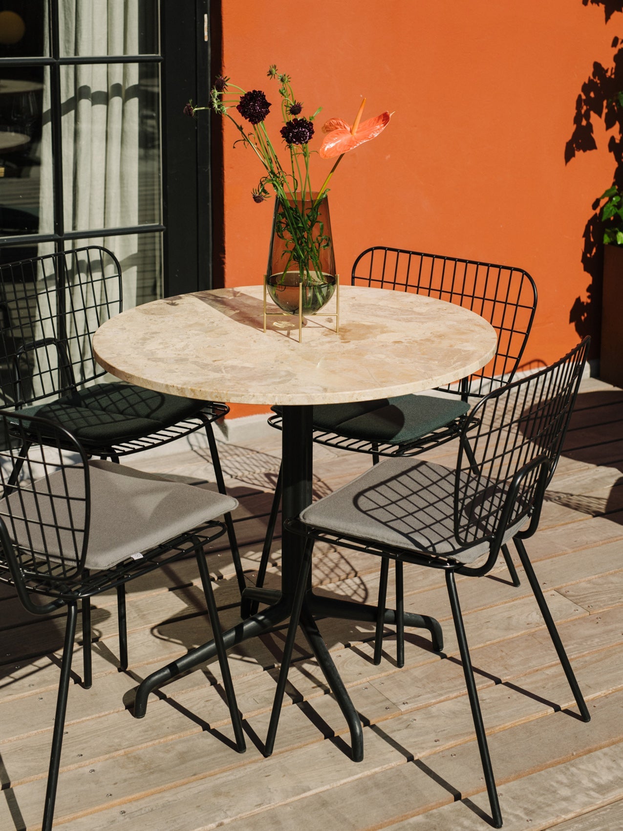 An outdoor patio scene featuring the Harbour Column Table with a marble top, four WM Dining Chairs, and an Echasse Vase.