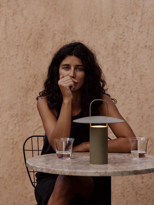 A woman with curly hair sits in a black WM String Dining Chair at a round marble Harbour Column Table with a green Ray Lamp.