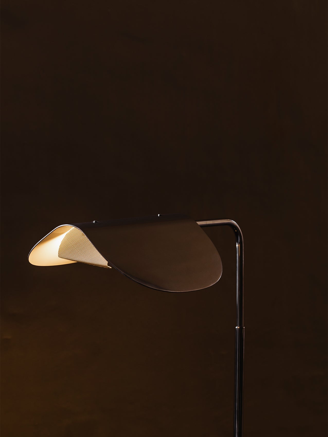 A close-up of the lit Wing Floor Lamp. Its polished aluminium, wing-like shade casts a warm glow against a dark background.