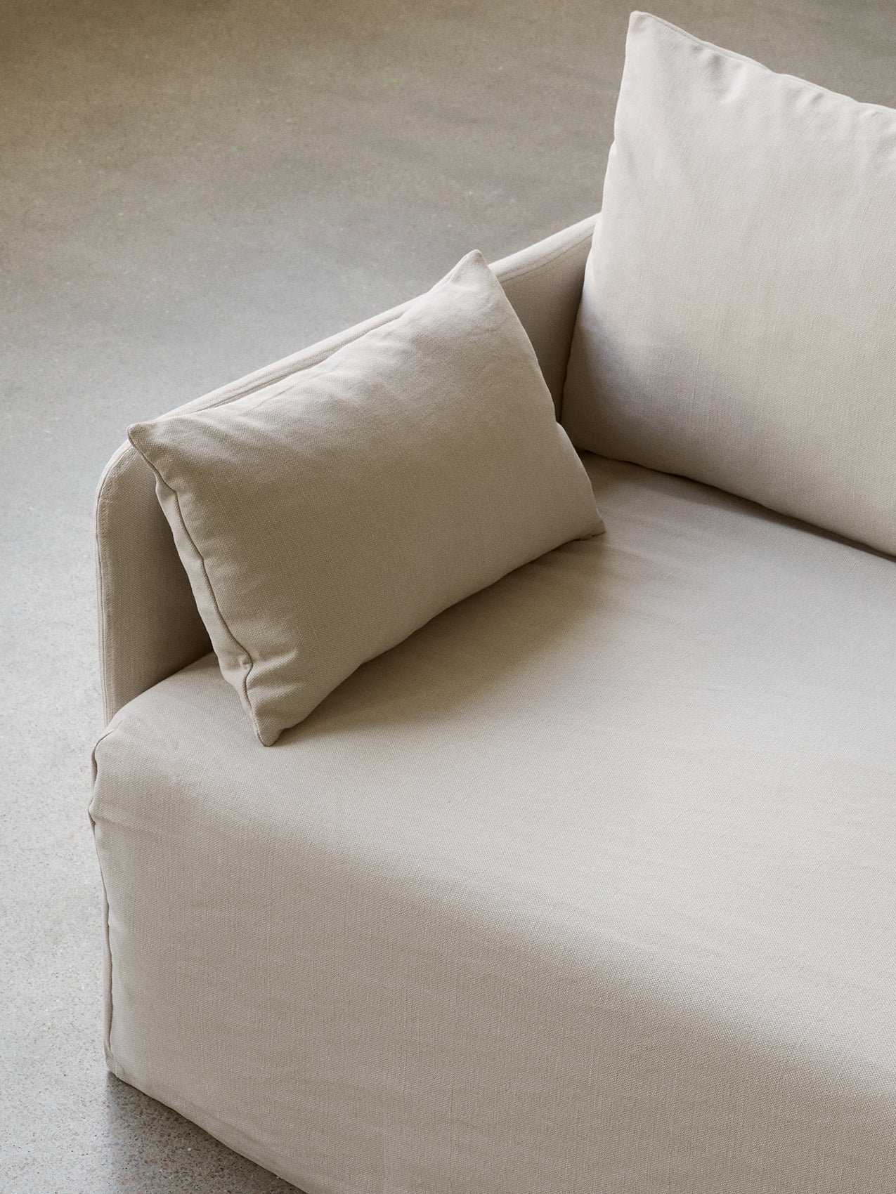 A close-up, high-angle shot of a sofa upholstered in a light beige, textured fabric, with soft cushions, on a concrete floor.