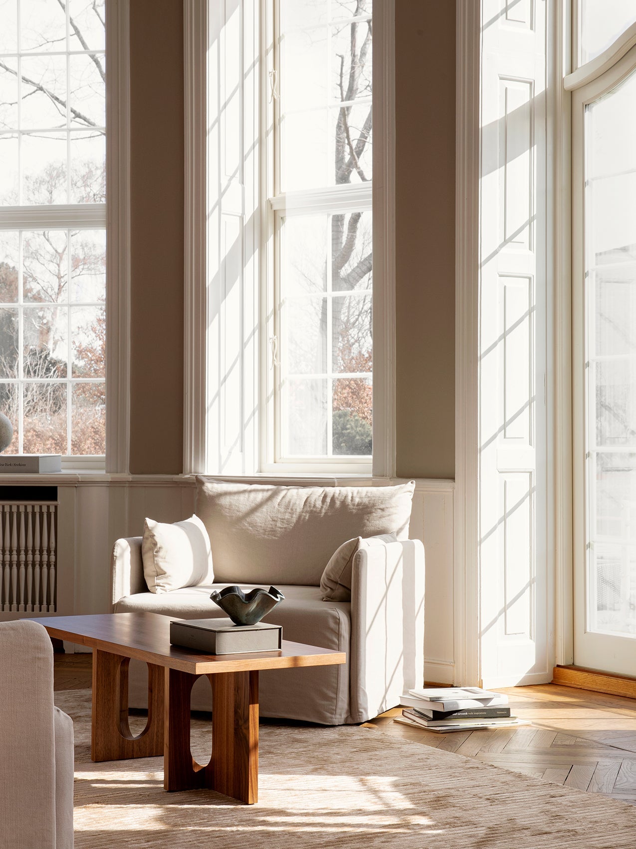A beige armchair and a solid wood coffee table in a sun-drenched room with large windows and a herringbone parquet floor.