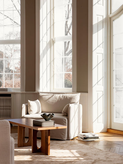 A beige armchair and a solid wood coffee table in a sun-drenched room with large windows and a herringbone parquet floor.